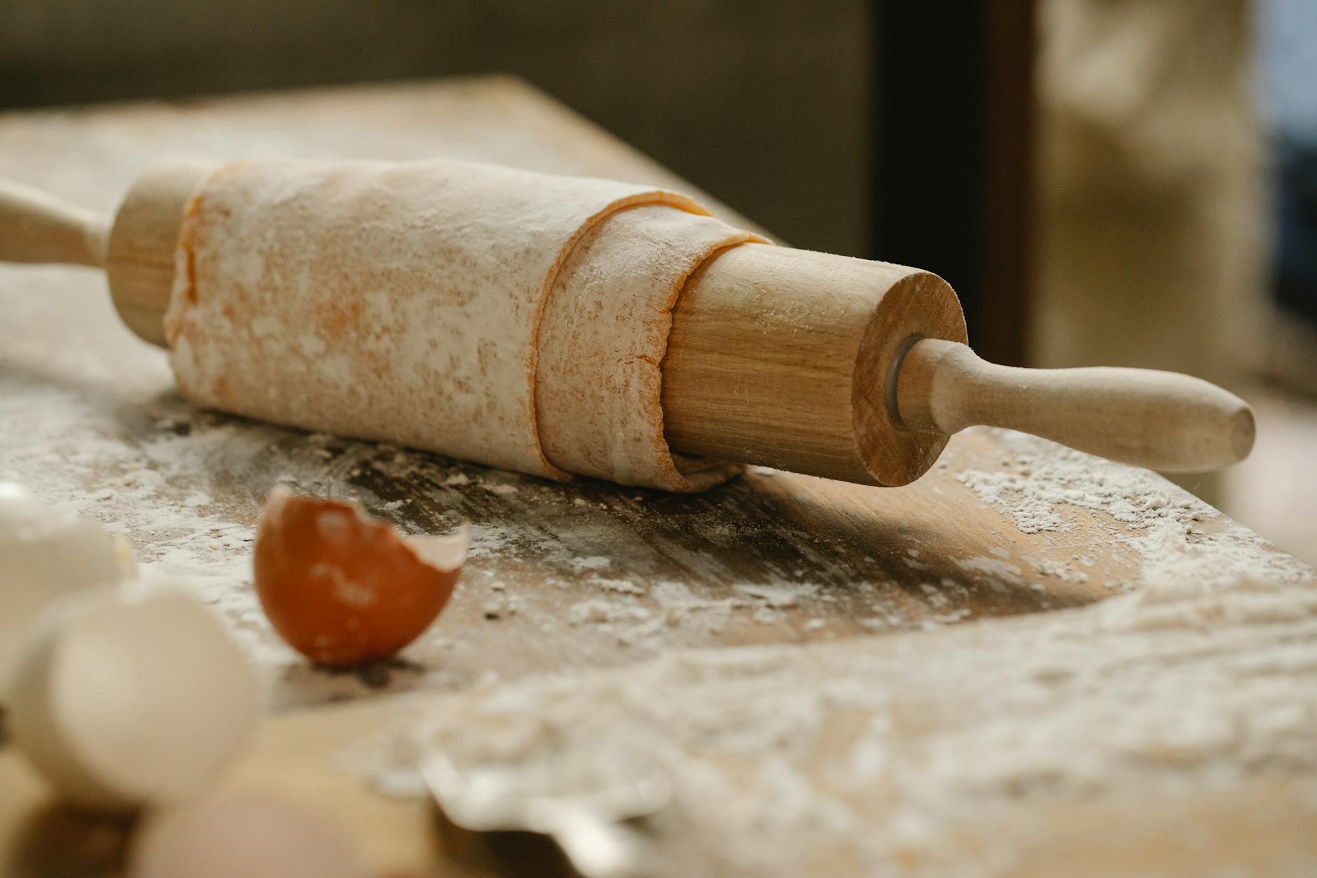 Rolling pin wrapped with uncooked sheet of dough placed on wooden table sprinkled with flour near eggshells in kitchen o